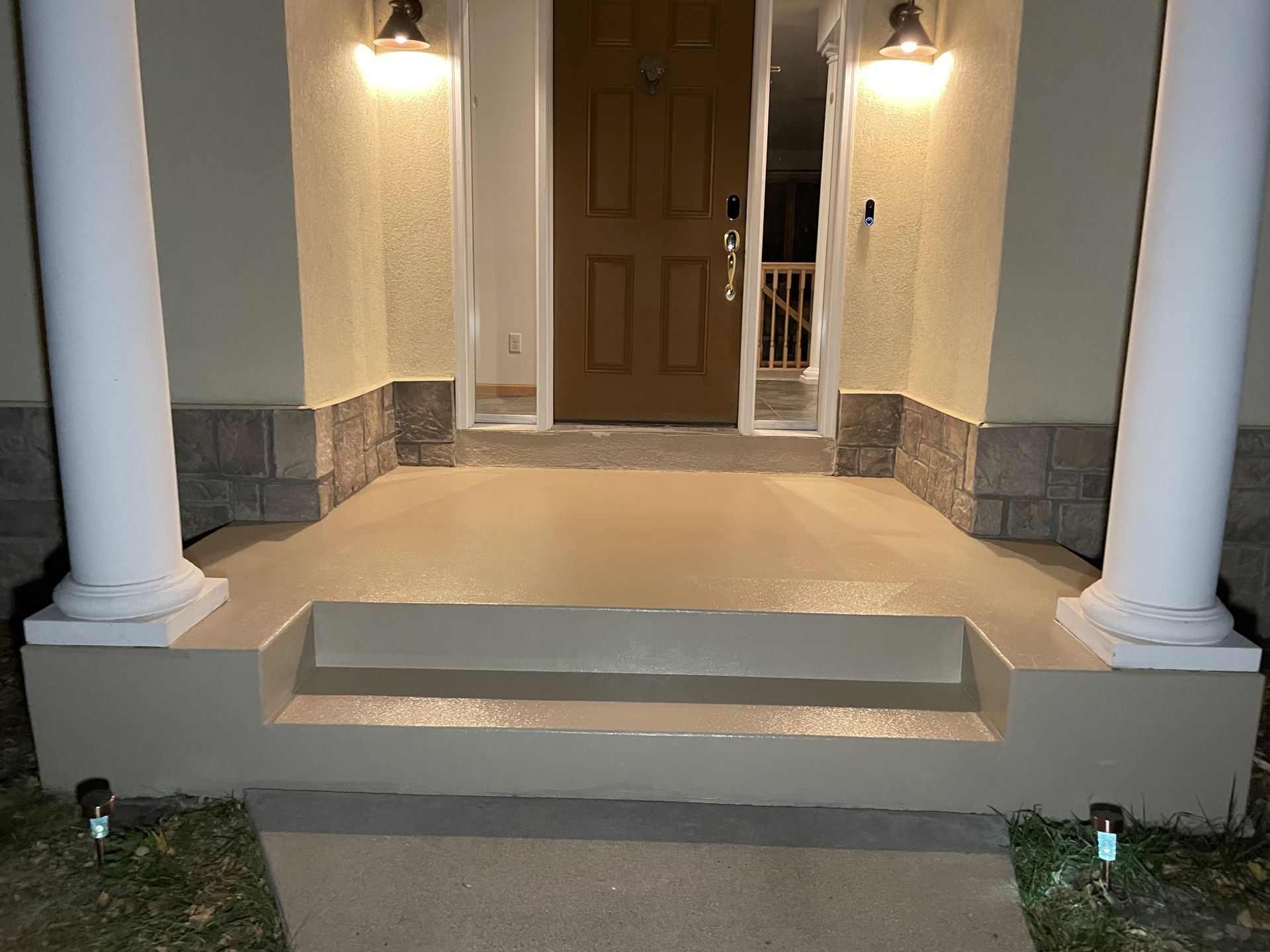 Front porch with tan steps, columns, and a brown door, lit by two wall-mounted lights.