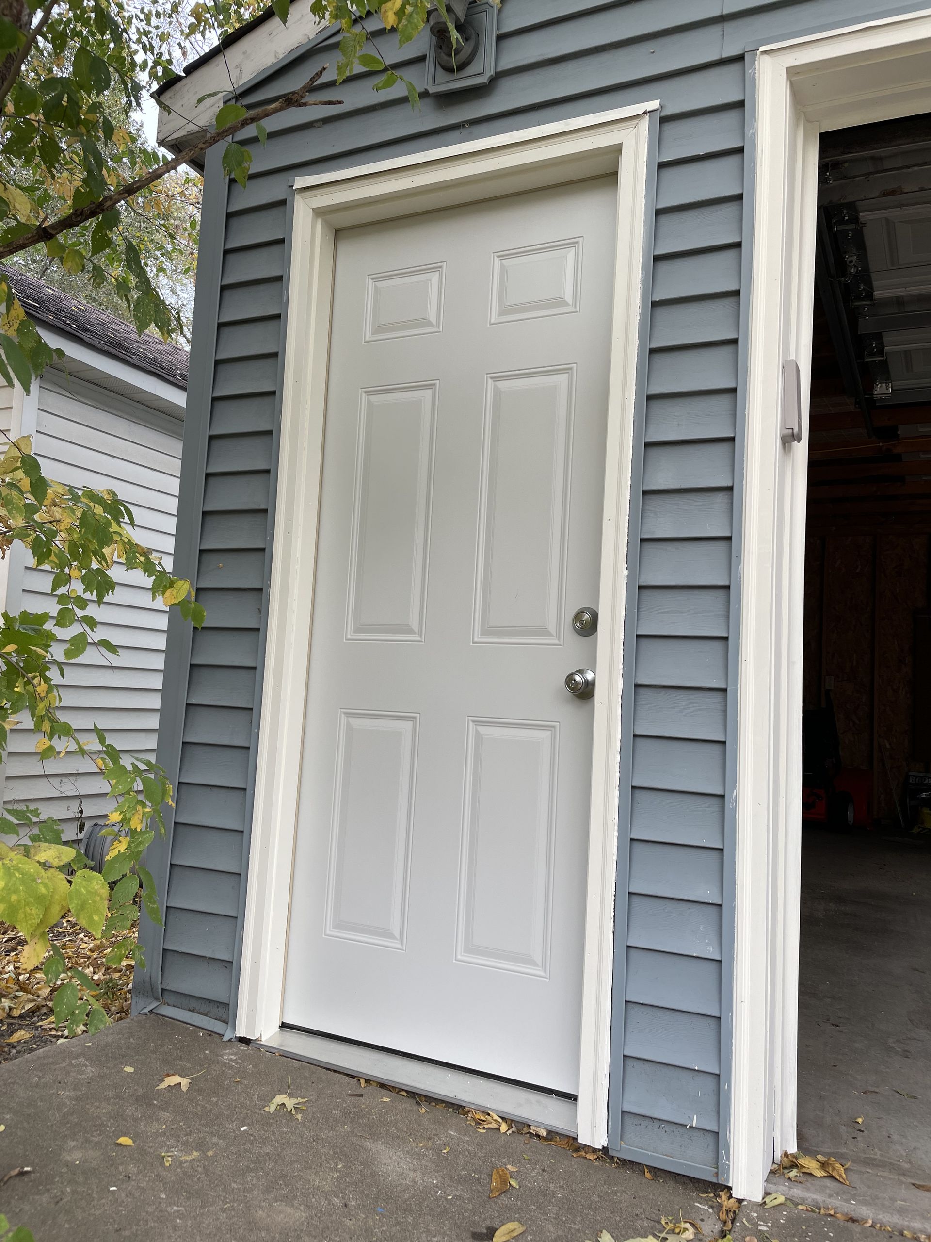 White door with white trim on a blue-gray house with a cement slab, next to an open garage.