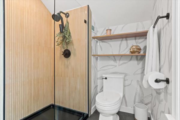 Small bathroom with a light wood shower stall, white toilet, and wallpaper.