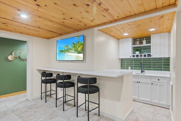 A finished basement with a bar area featuring seating, a TV, white cabinetry, and a wooden ceiling.