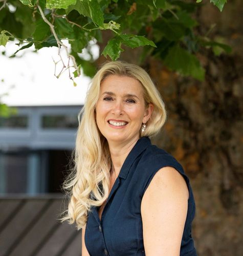 Smiling woman with long blonde hair, wearing a navy sleeveless top, sitting outdoors under a tree. Used for the bio section on The Unconventional You's About page.