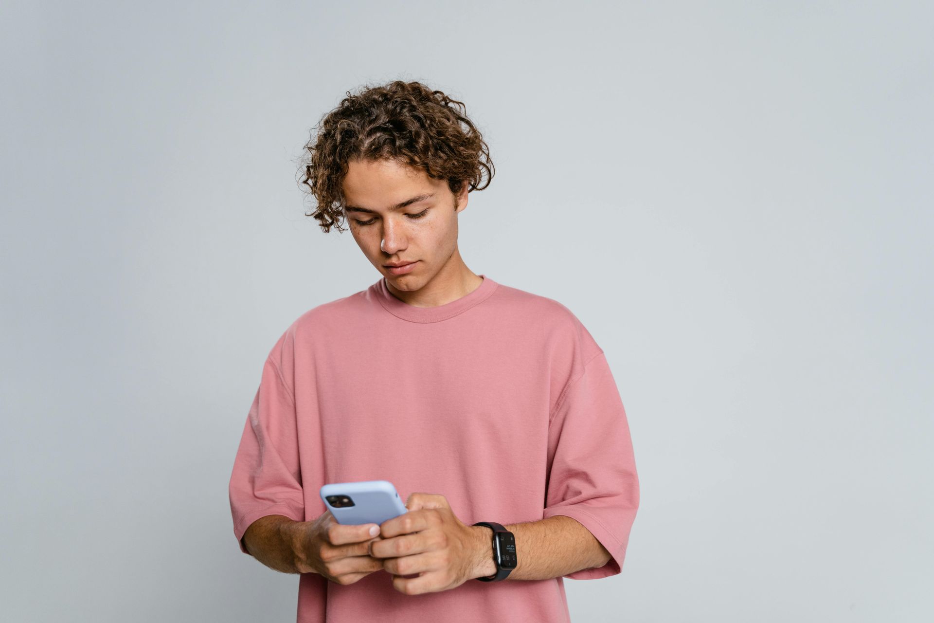 A teen wearing a pink t-shirt, looking at their smartphone against a neutral background