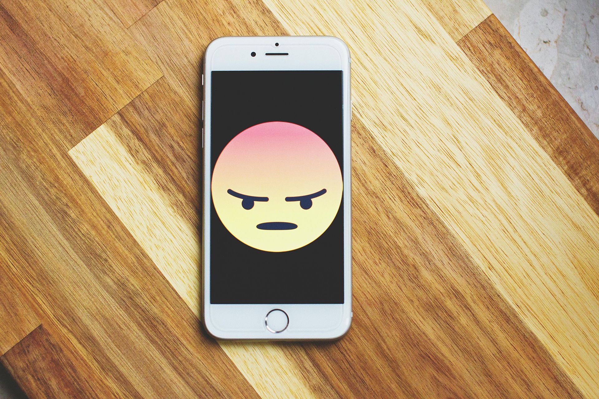 A smartphone placed on a wooden surface displays an angry emoji with a red-to-yellow gradient face a