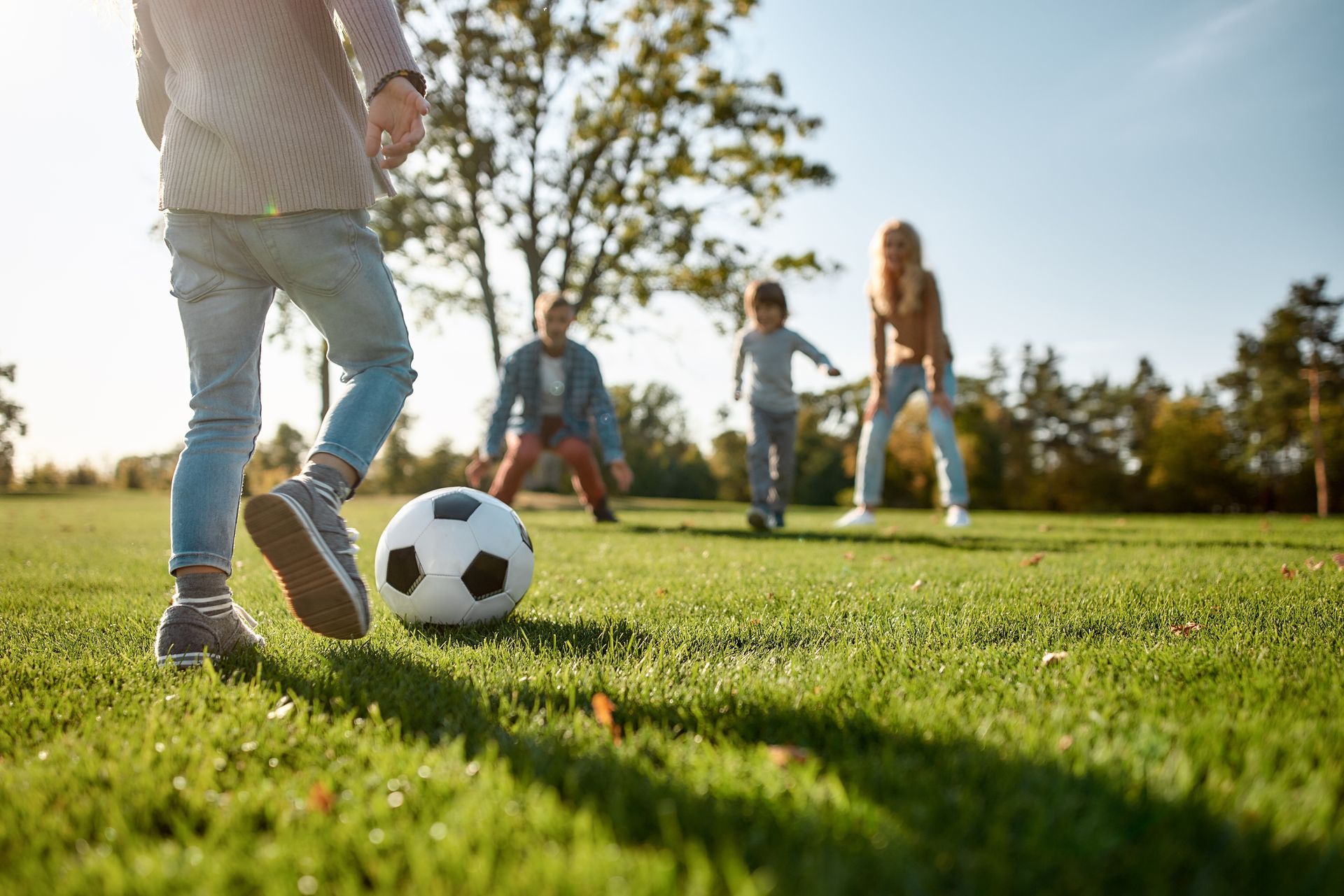 Child about to kick soccer ball with family in the background on a sunny grassy field.