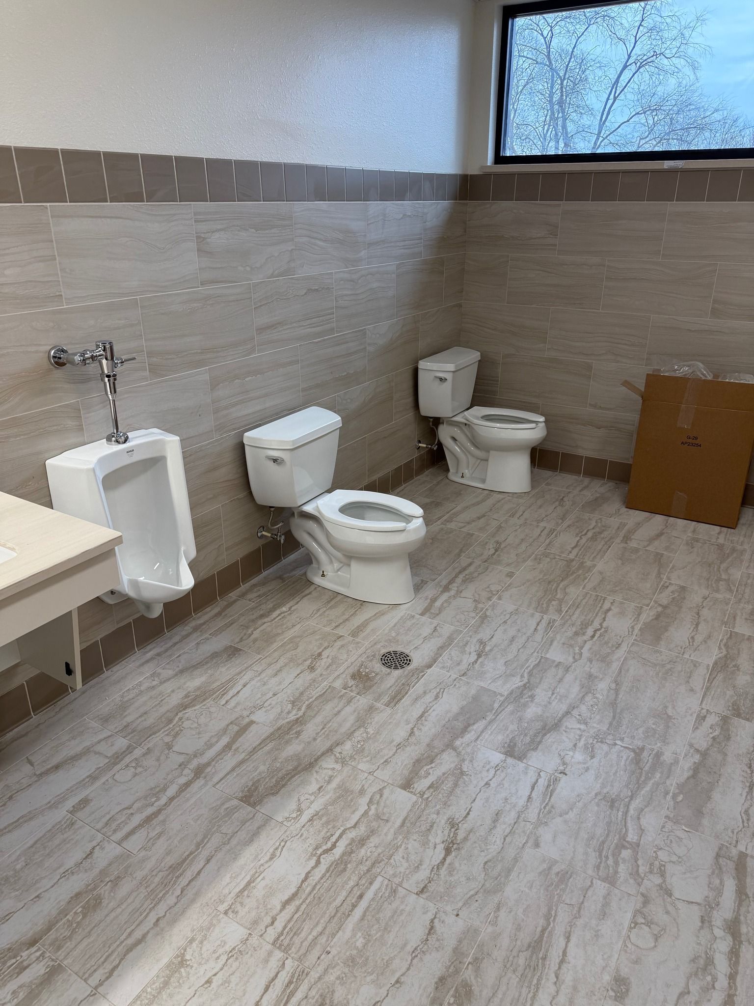 Public restroom with urinals, toilets, and a large frosted window on tiled walls and floor.