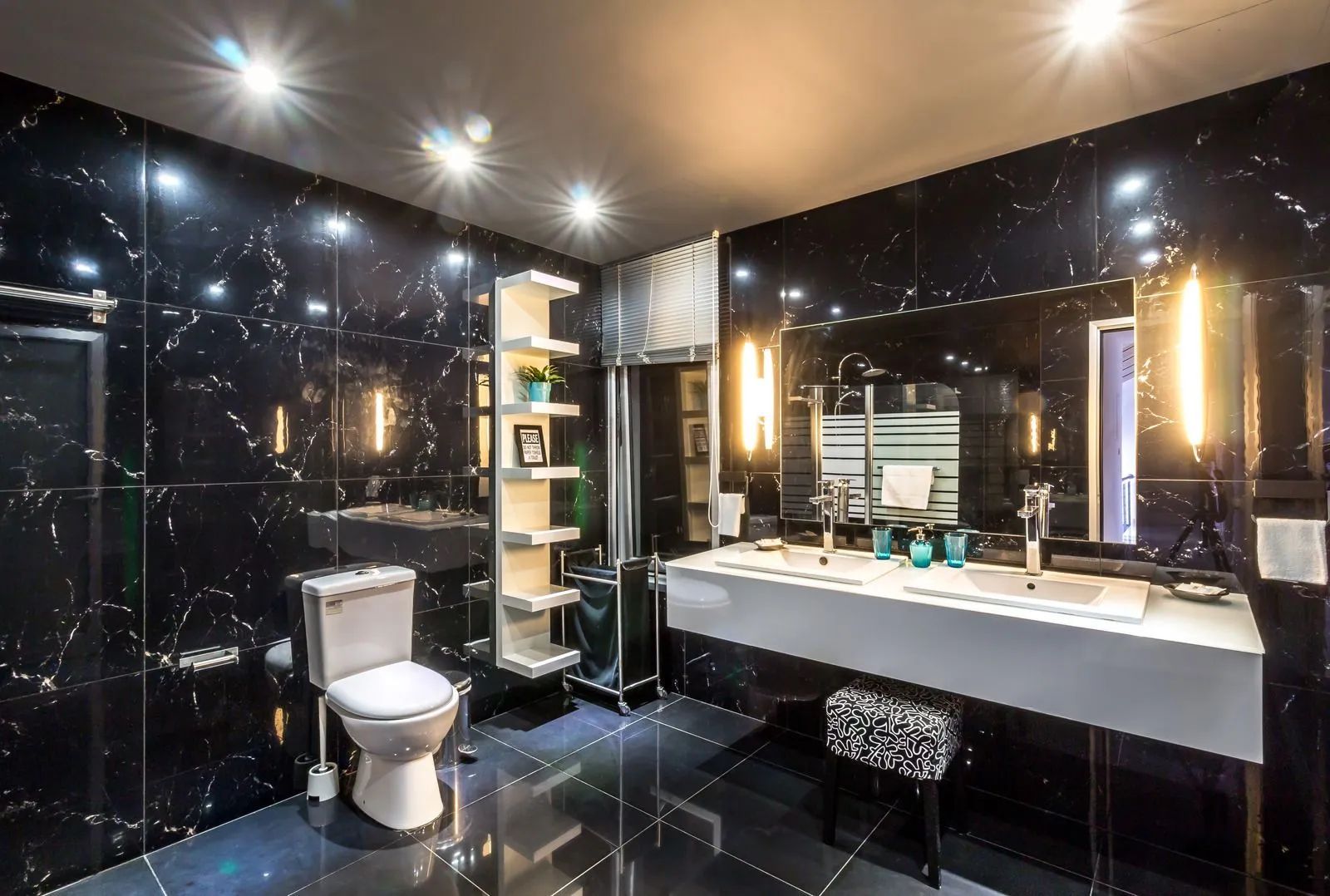 Modern black-tiled bathroom with toilet, vanity sink, lit mirrors, and shelving