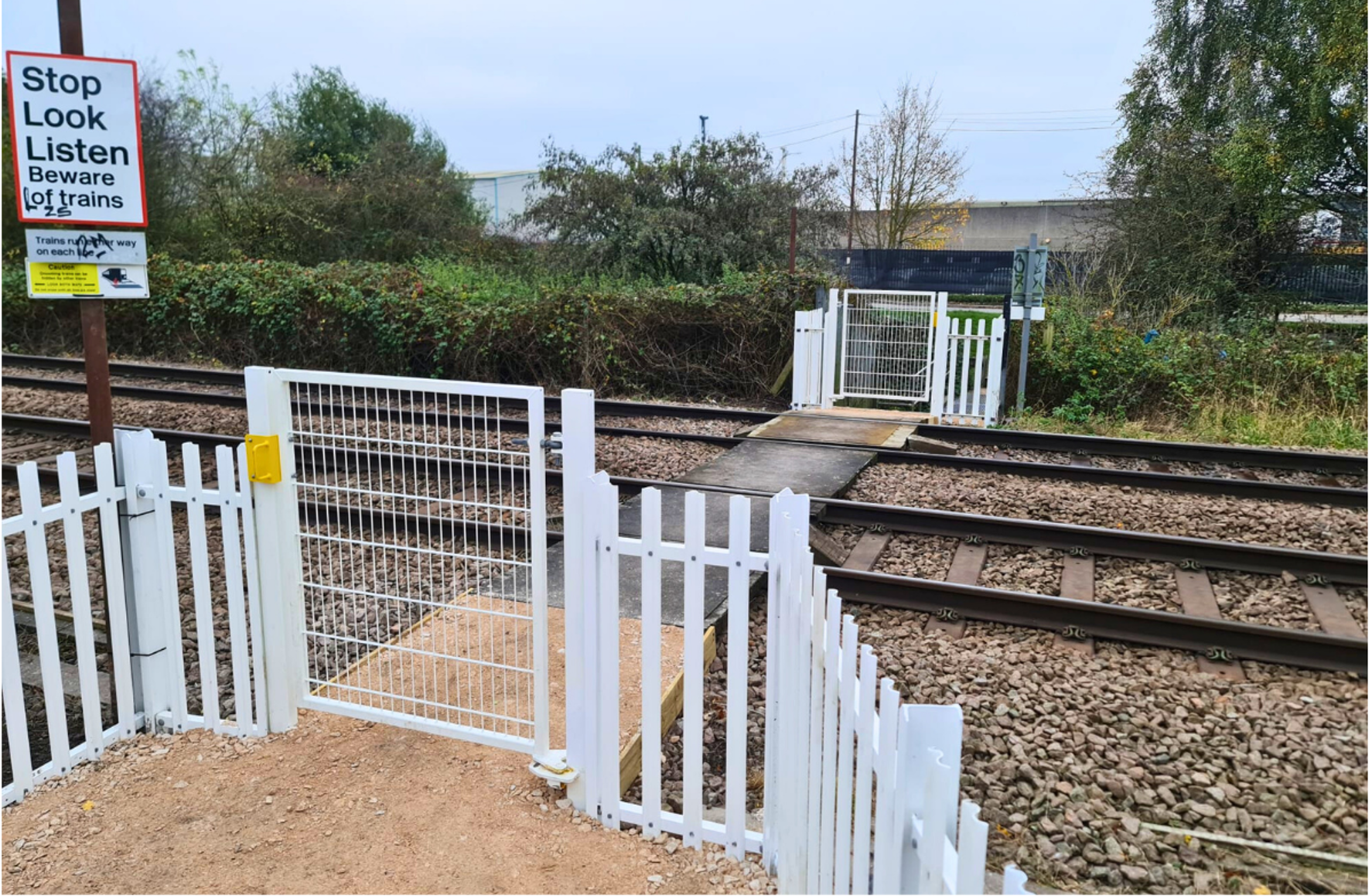 White fenced railway crossing with gates, warning sign, and tracks under cloudy sky