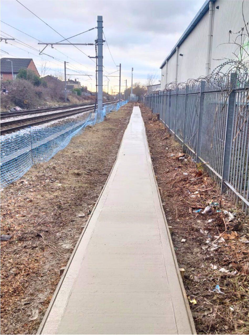 Narrow concrete walkway beside railroad tracks and a chain-link fence under a cloudy sky