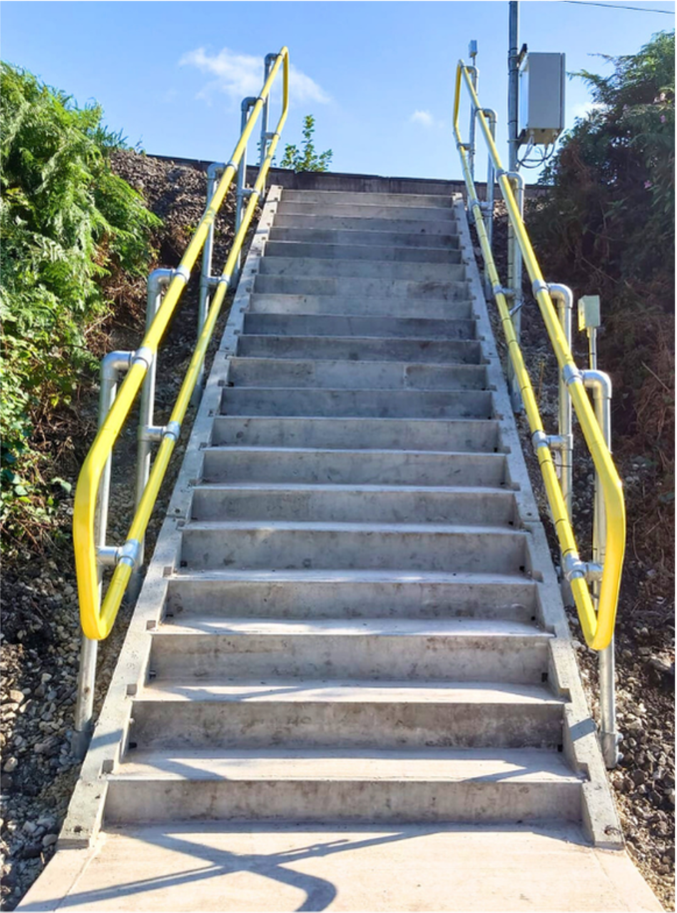 Concrete outdoor staircase with yellow handrails leading uphill between shrubs and buildings