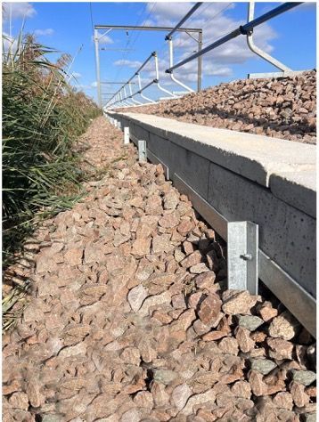 Rocks piled along a rail line under a clear blue sky, with a raised concrete track and metal railing.