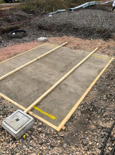 Rectangular gravel pad marked with wooden framing and a small concrete utility box in the foreground.