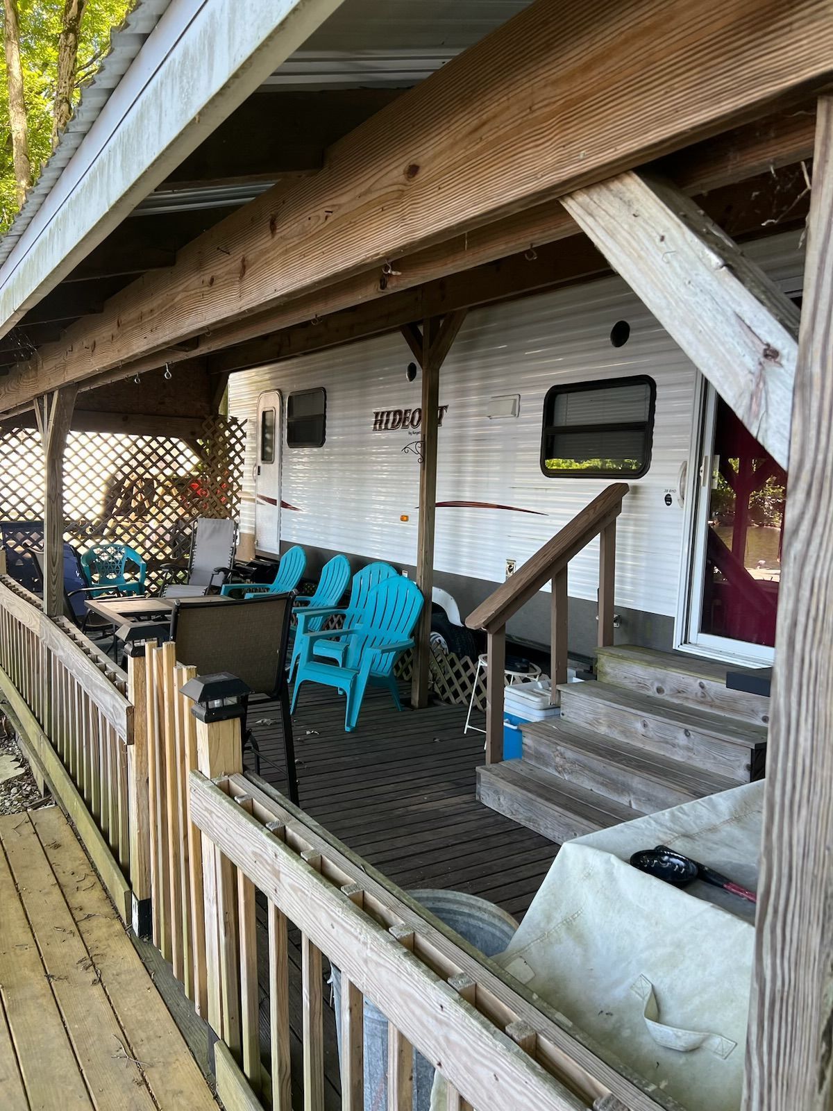 A rv is parked under a covered porch with blue chairs