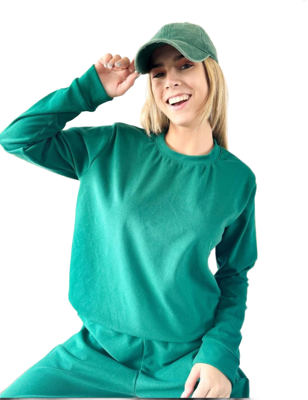 Mujer con sudadera verde y gorra de béisbol sonríe, con la mano en la gorra.