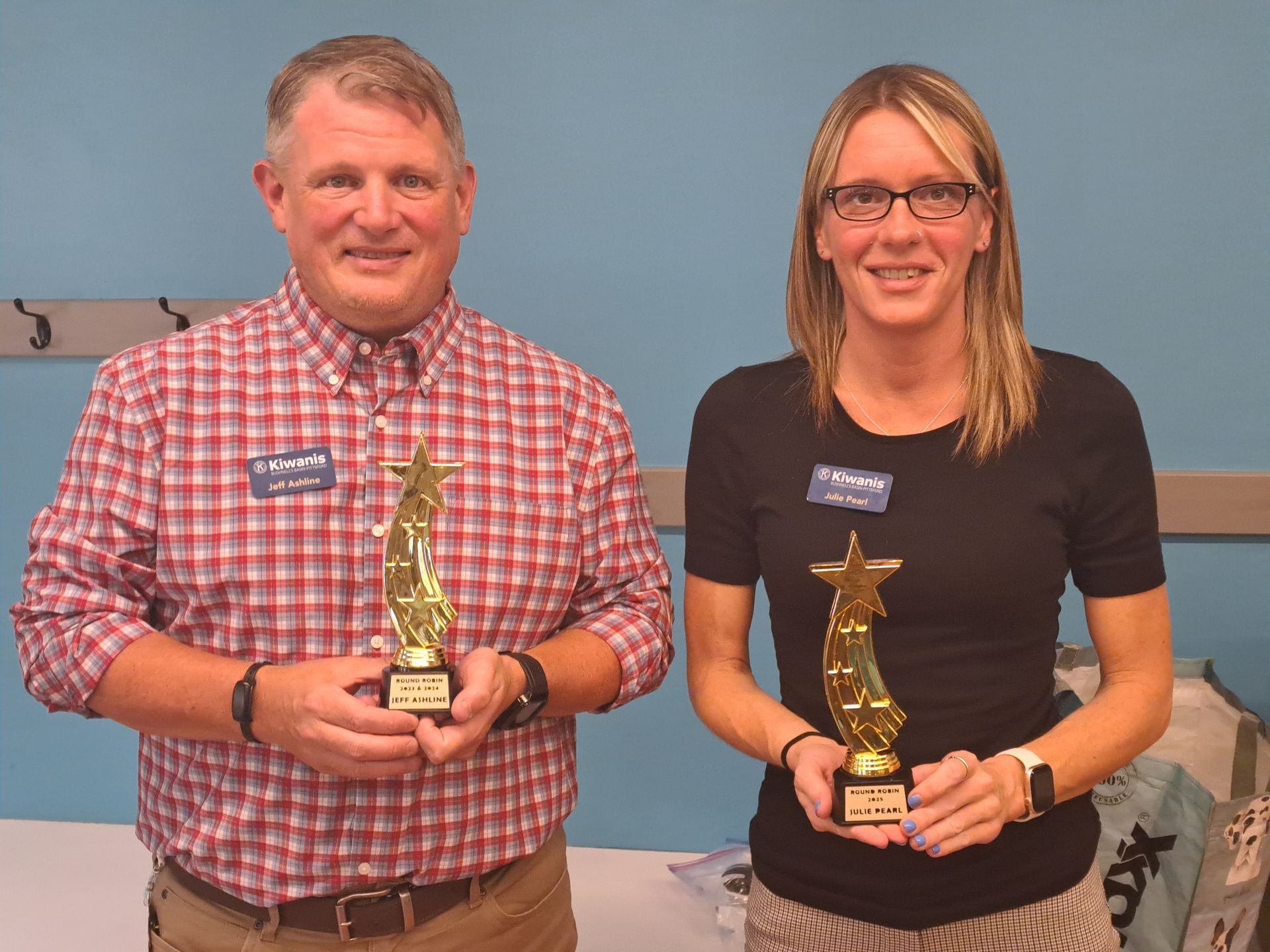 Man and woman holding star-shaped trophies, smiling. The man wears a plaid shirt; the woman wears glasses and a black shirt.