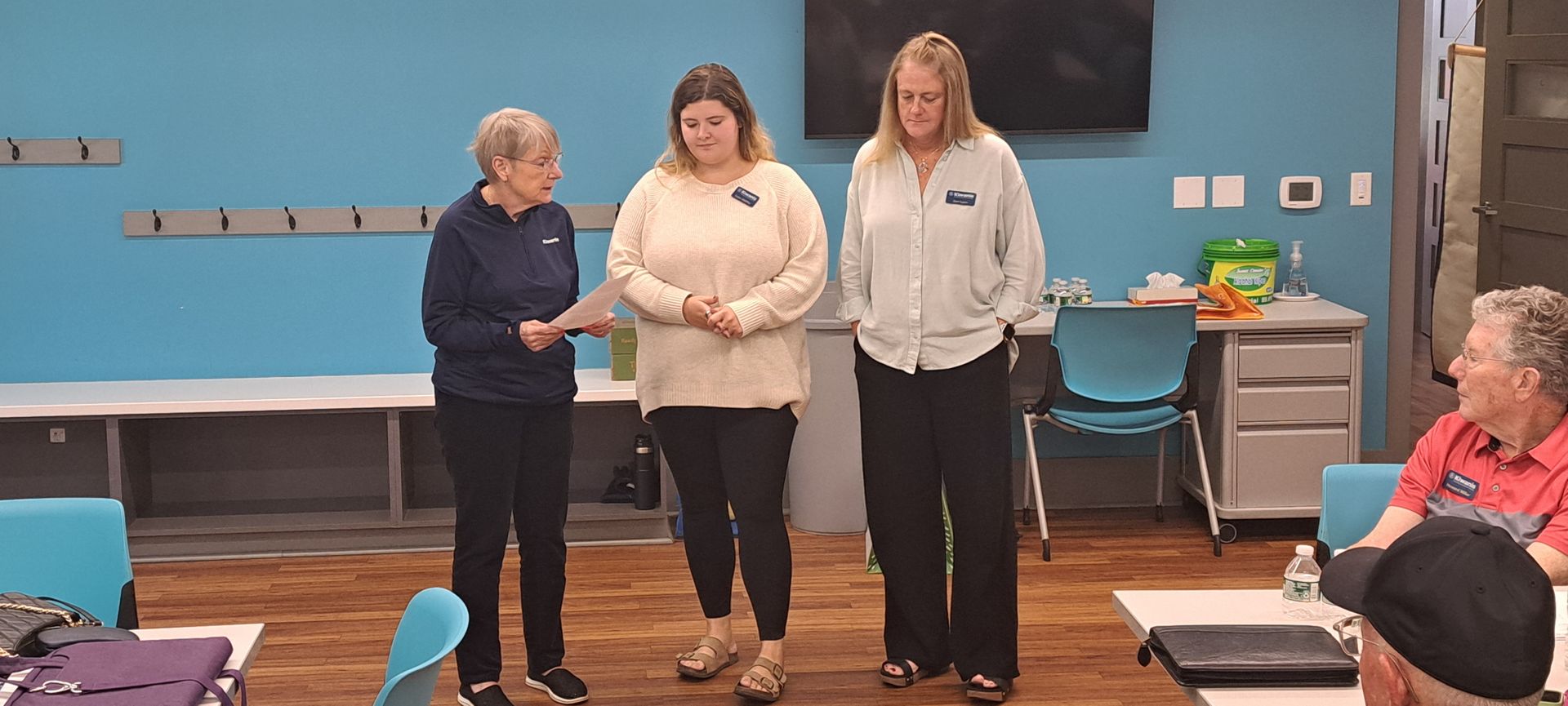 Three people stand in a room, two facing forward. Another person is seated, facing the others.