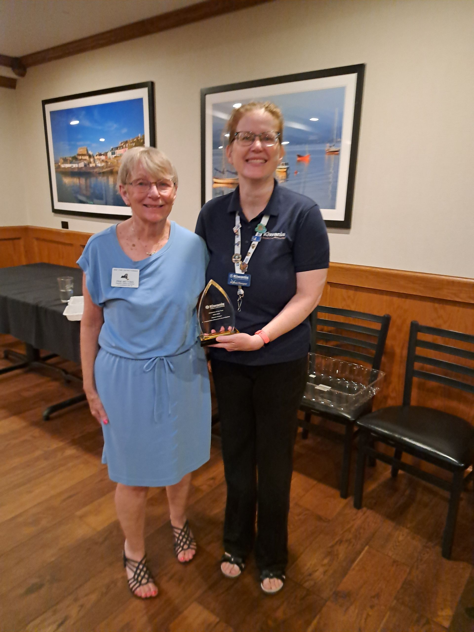 Two women holding an award, smiling, indoors. One in blue dress, other in uniform. Background: framed artwork, chairs.