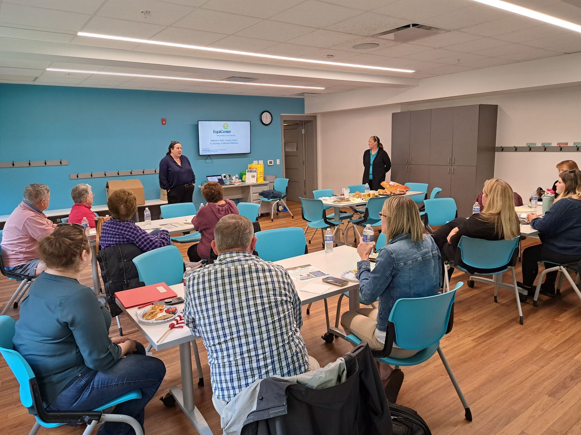 People in a workshop, two presenters, tables, teal chairs, blue wall, white ceiling, and a wooden floor.