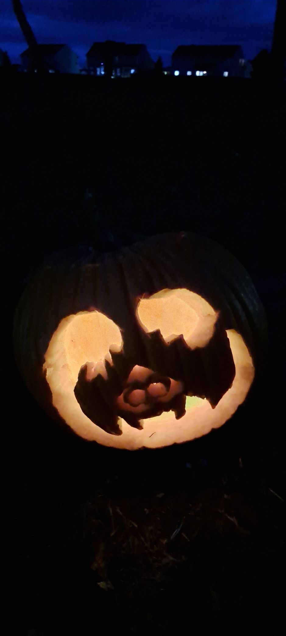 Lit-up jack-o'-lantern with a carved face, in a dark setting, under a night sky with house lights in the background.