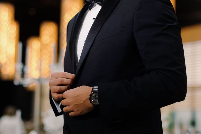 Man in a black tuxedo adjusting his cufflinks, with warm blurred lights in the background