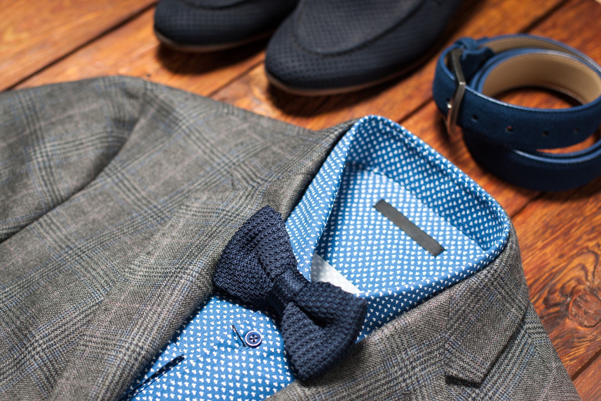 Brown tweed suit jacket with blue dress shirt and black bow tie beside dress shoes and belt on wooden floor
