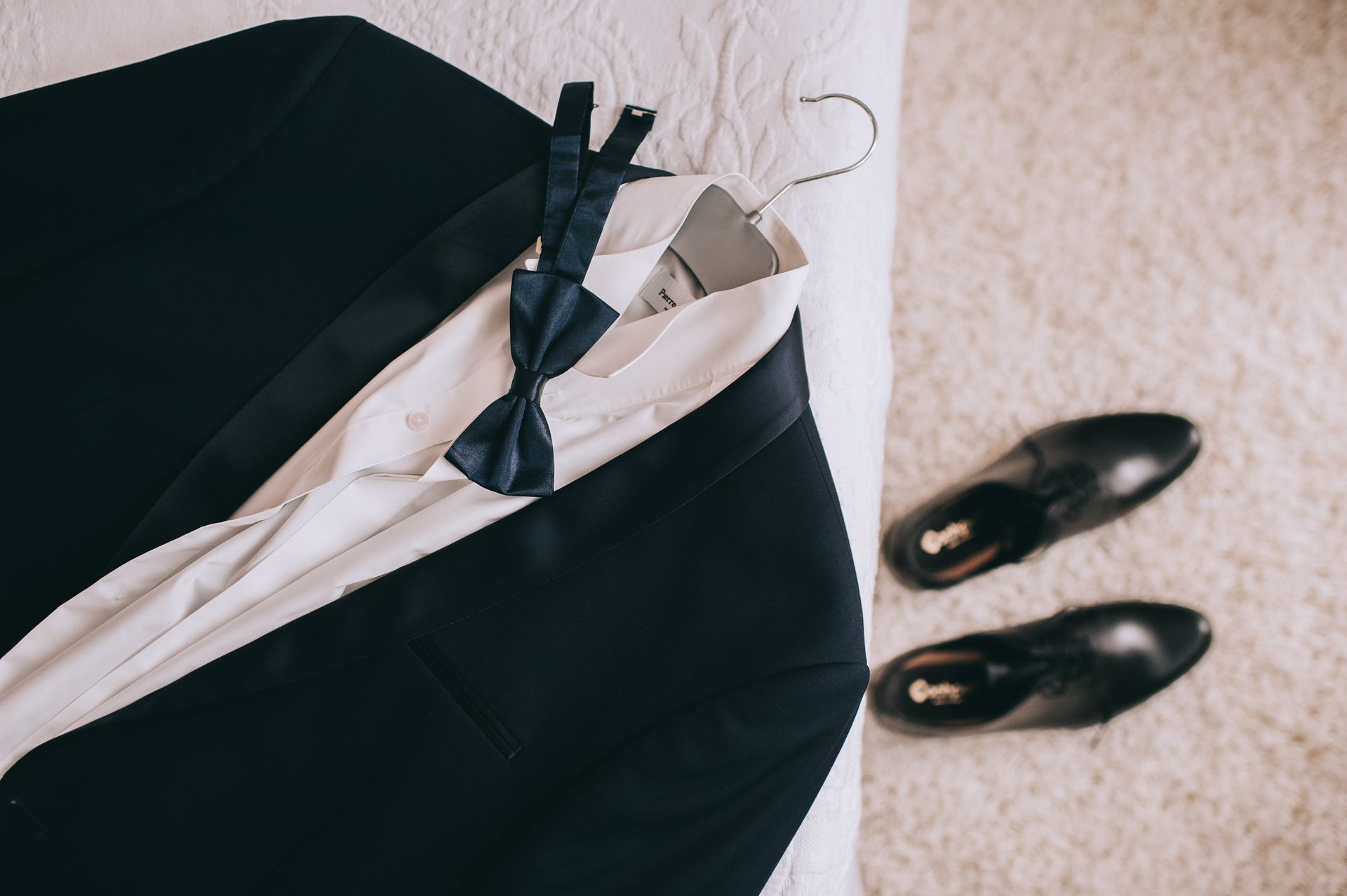 Black suit jacket with white shirt and dark tie on a hanger, with black shoes on carpet nearby