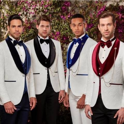 Four men in white tuxedos with colored lapels pose in front of a floral wall.