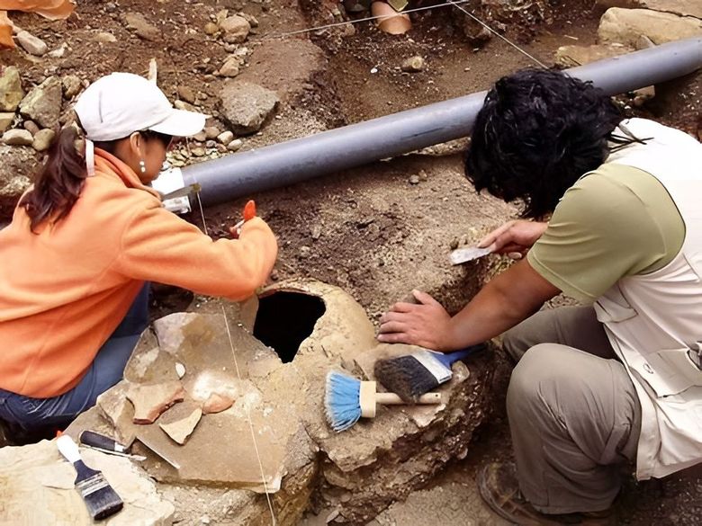 Dos trabajadores limpian una excavación cubierta de tierra cerca de una tubería negra, utilizando cepillos y herramientas.