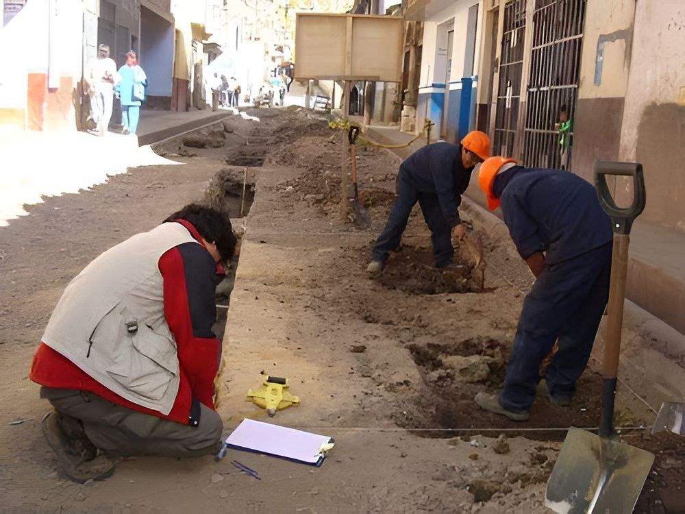 Trabajadores cavando una zanja en una obra de construcción, con una persona arrodillada en primer plano.