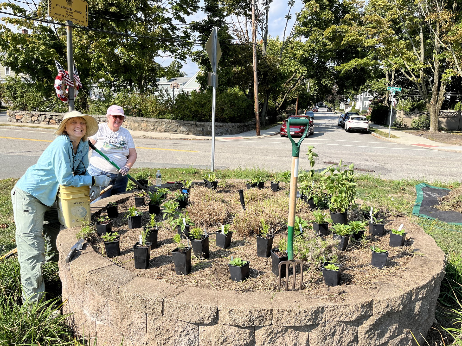 A raised brick circular garden bed, with many small seedlings laid out for planting. Two gardeners smile toward the camera.