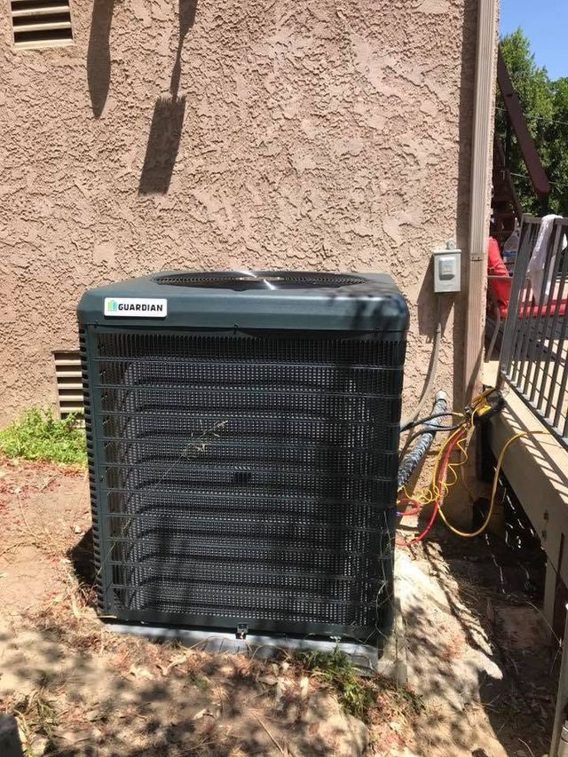 Outdoor HVAC condenser unit beside a stucco wall with service wires and a fence gate nearby