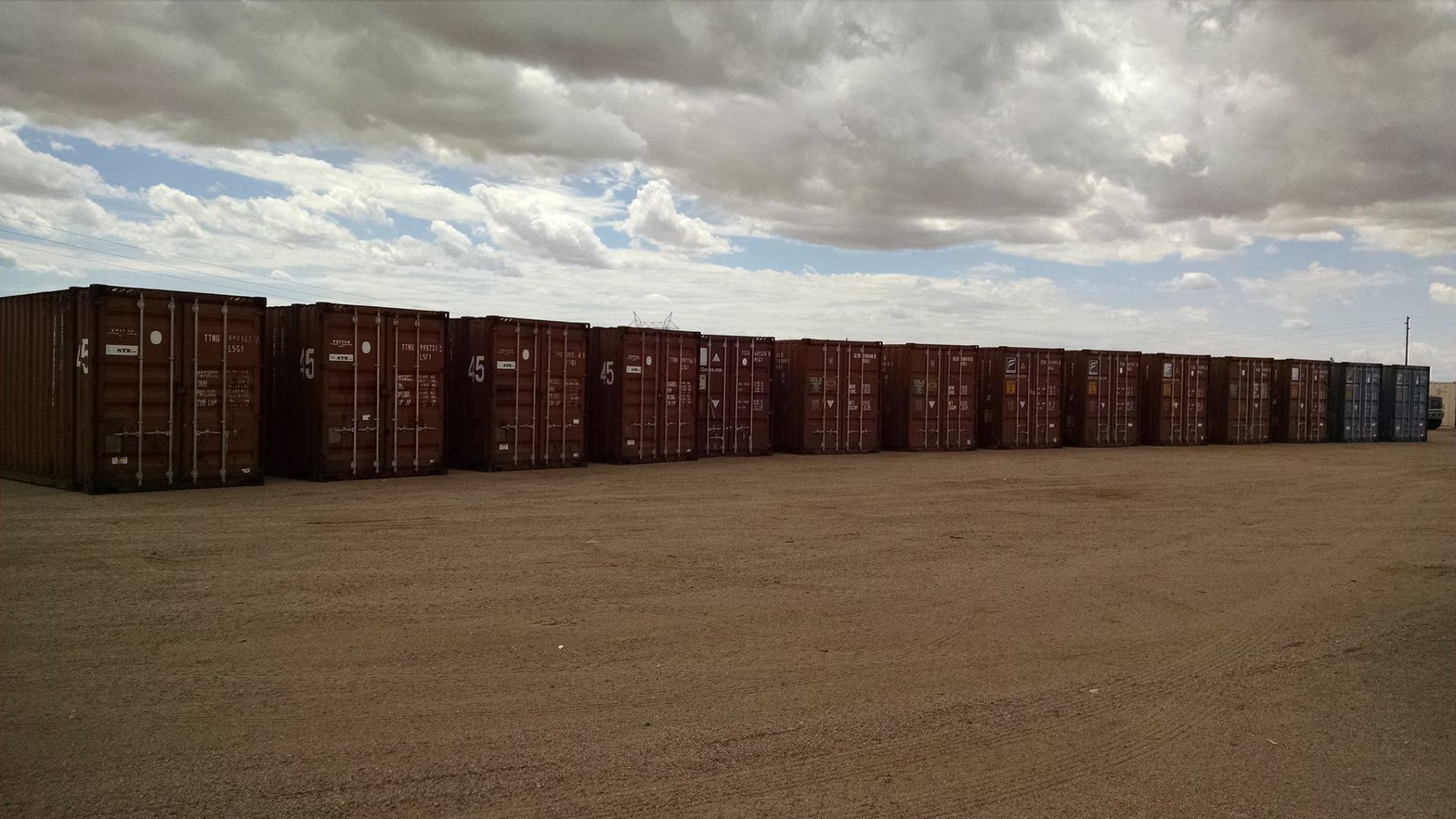 A row of shipping containers are lined up in a dirt field