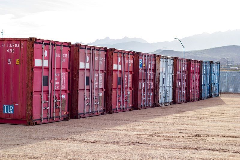 A row of shipping containers are lined up in a dirt field.