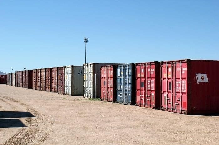 A row of shipping containers are lined up in a dirt lot
