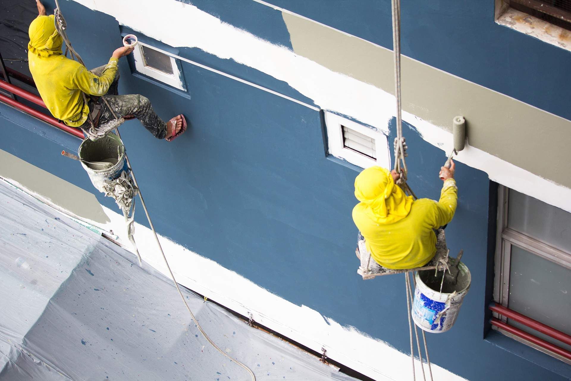 two men are painting a building with buckets attached to ropes .