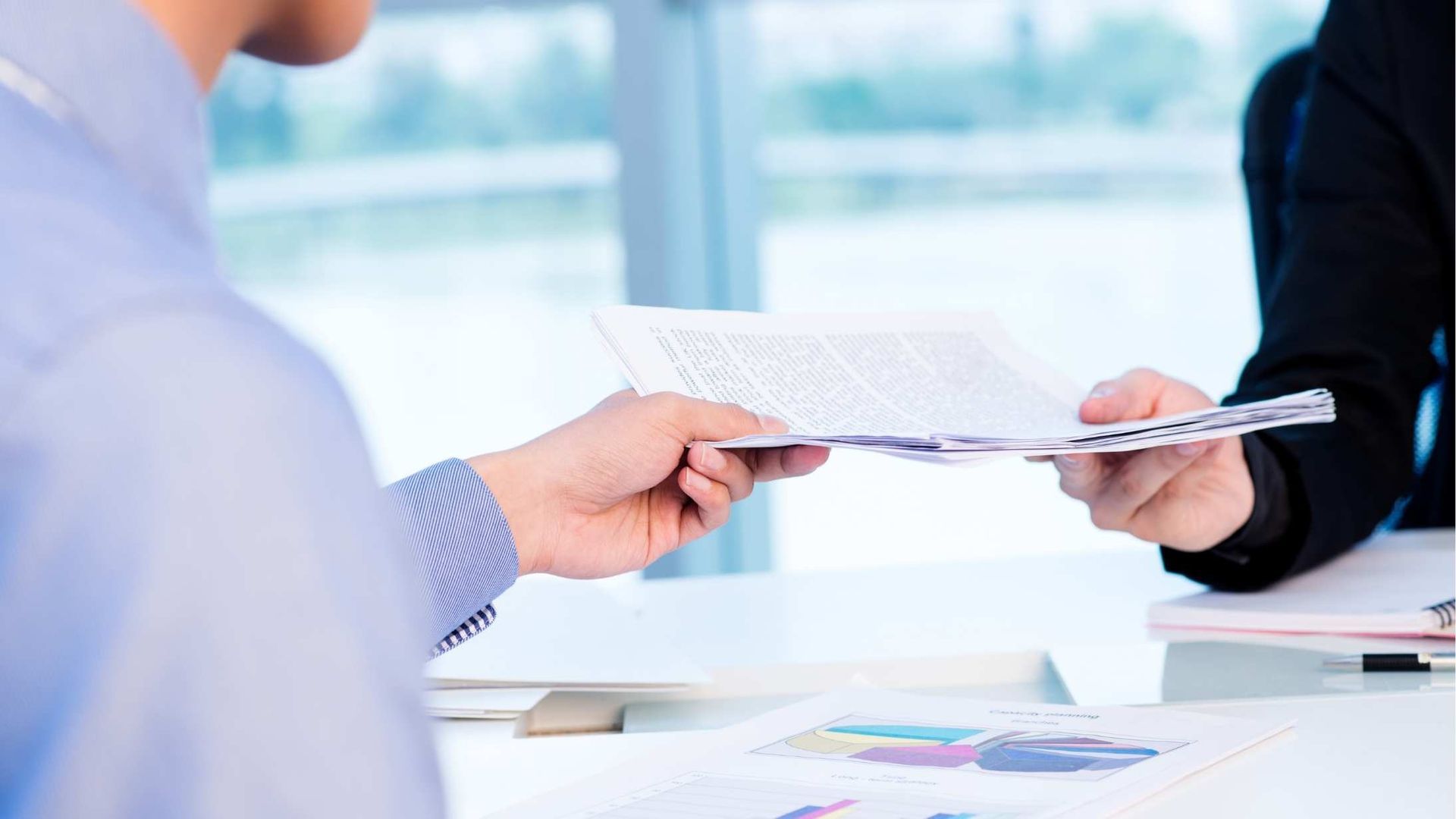 Two people exchange printed documents across a desk in a bright office setting.