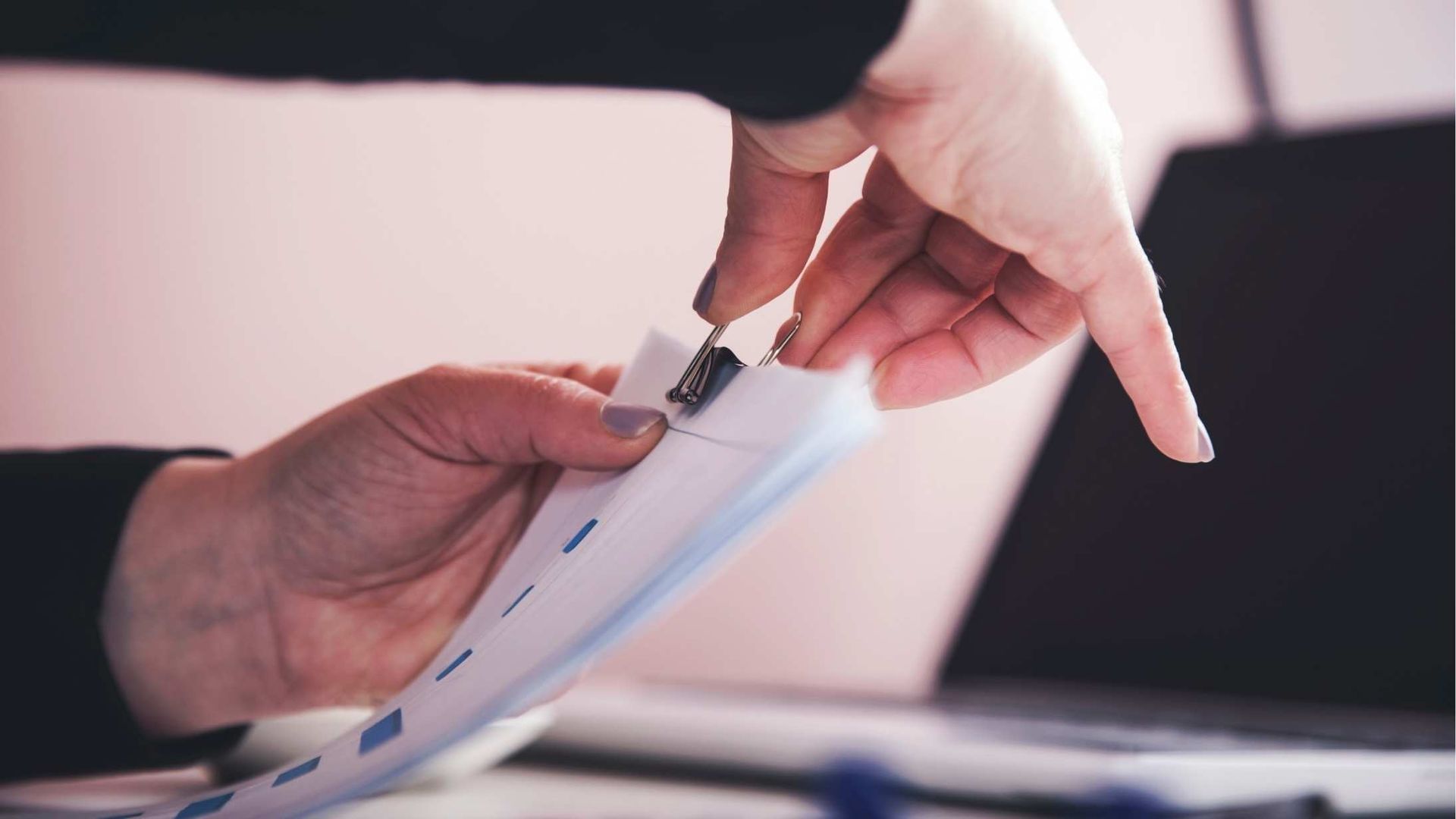 Hands passing a document at a desk, with a laptop in the background.