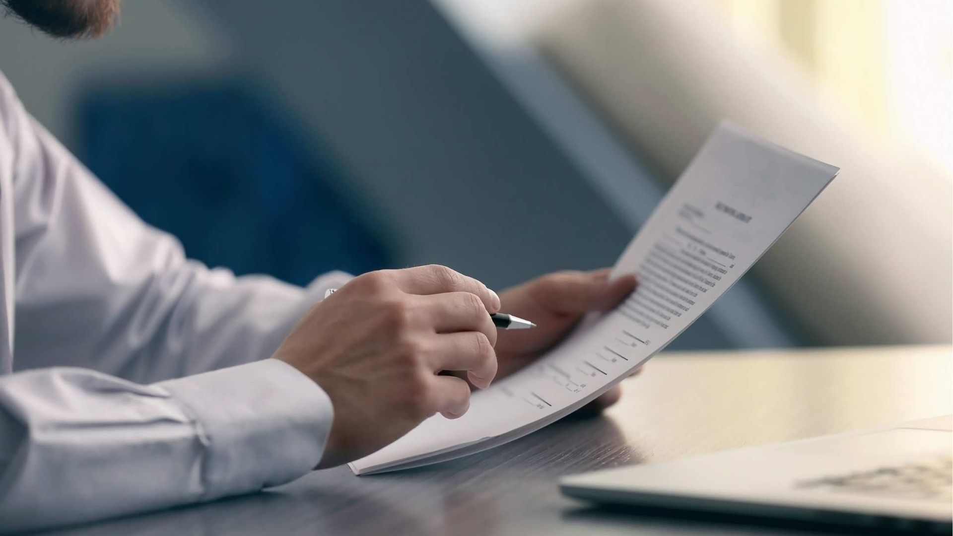 Close-up of hands reviewing and marking a document at a desk with a laptop nearby