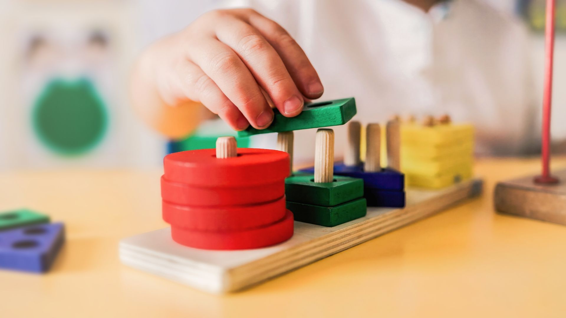 Child's hand placing green block on peg toy with red, blue, and yellow shapes.