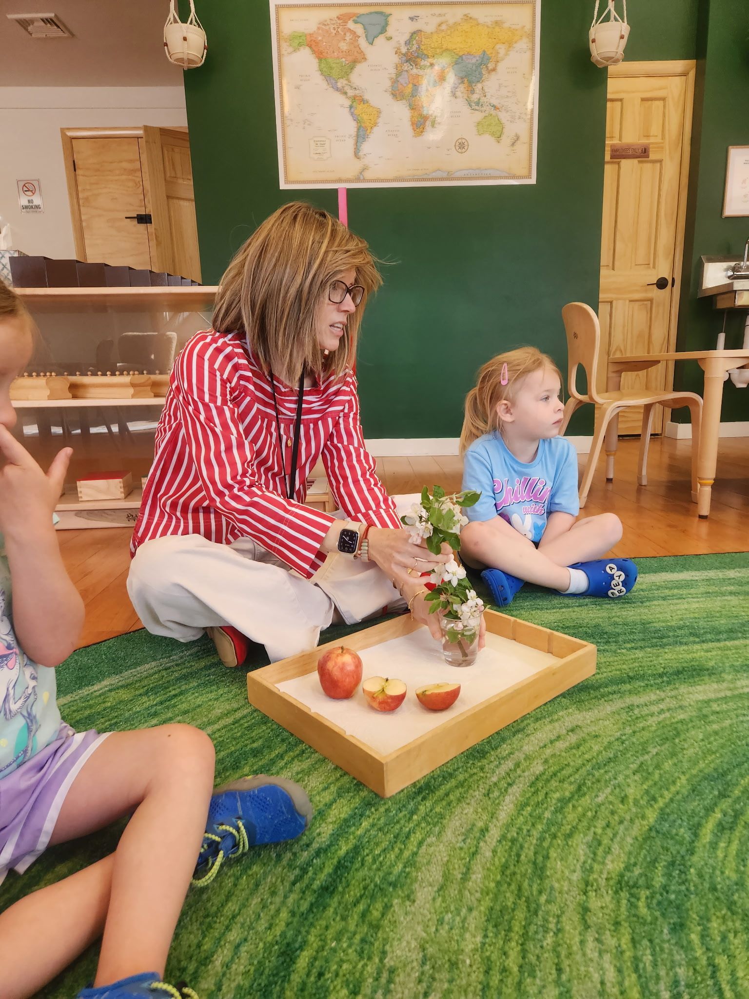 Teacher with two children on a green rug, looking at peaches and flowers on a tray. A world map hangs in the background.