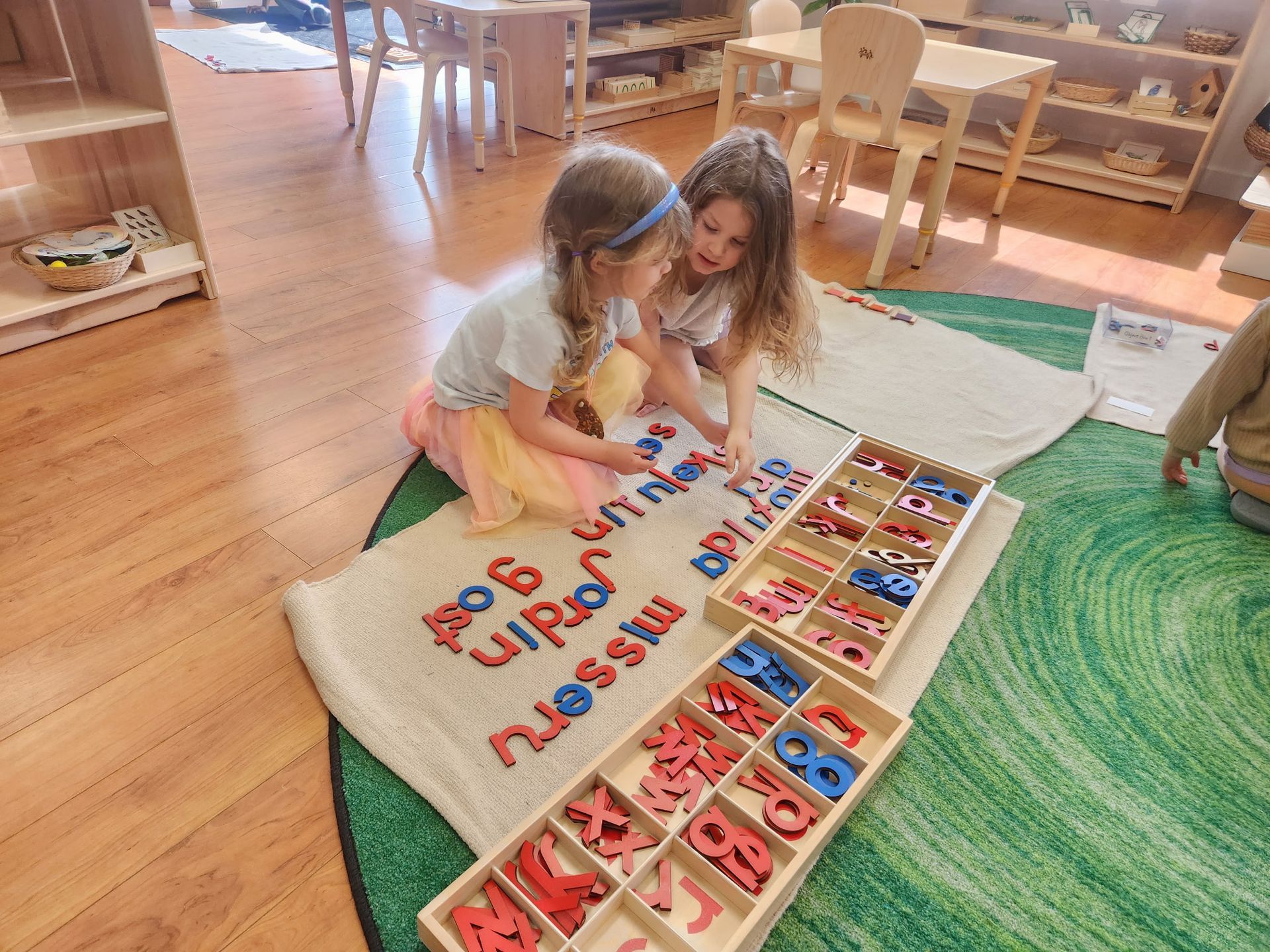 Two children working with letter tiles on a rug in a classroom.