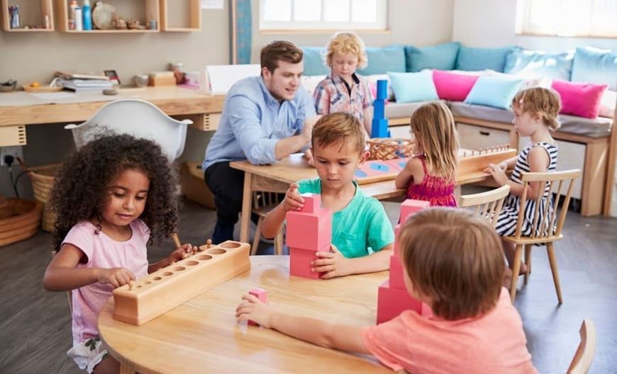 Children and a teacher at a table working with blocks and other educational toys in a classroom.