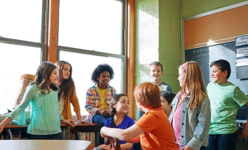 Children chatting in a sunlit classroom near a large window, some sitting on a desk.