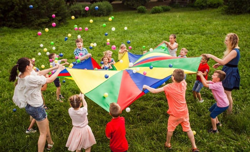 Children and adults playing with a colorful parachute and balls on a grassy field.