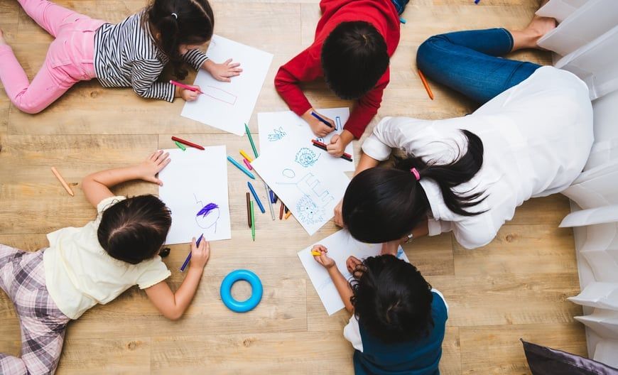 Children and a person drawing on paper together on the floor with colored pencils and a blue ring.