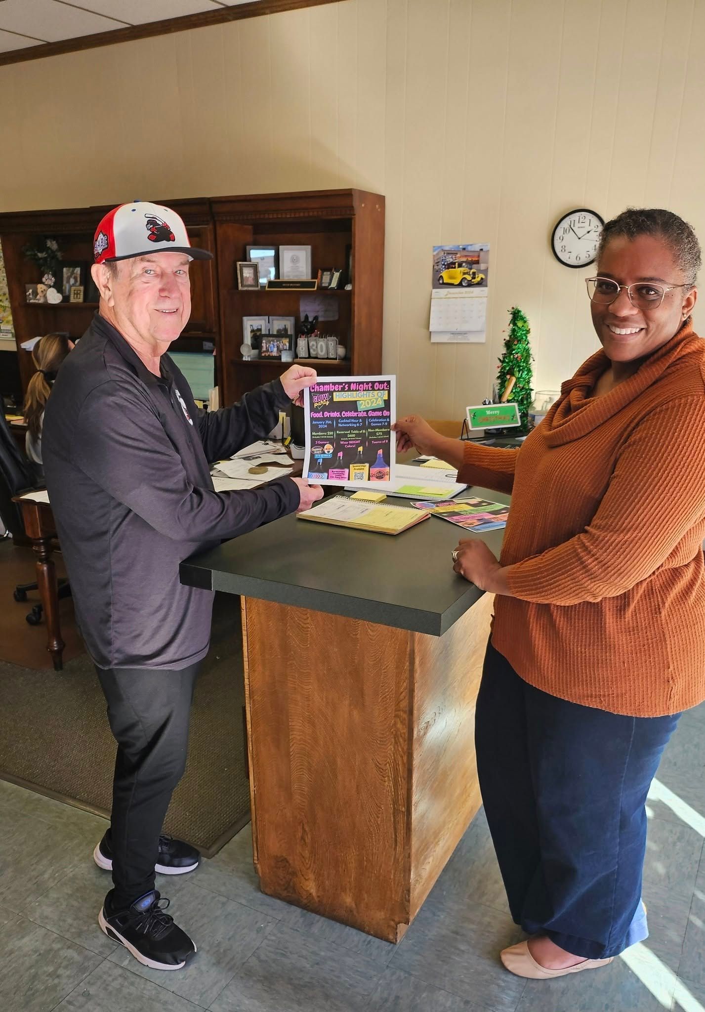 Two people at a desk, looking at a flyer. One wears a baseball cap, the other a sweater. Indoors.