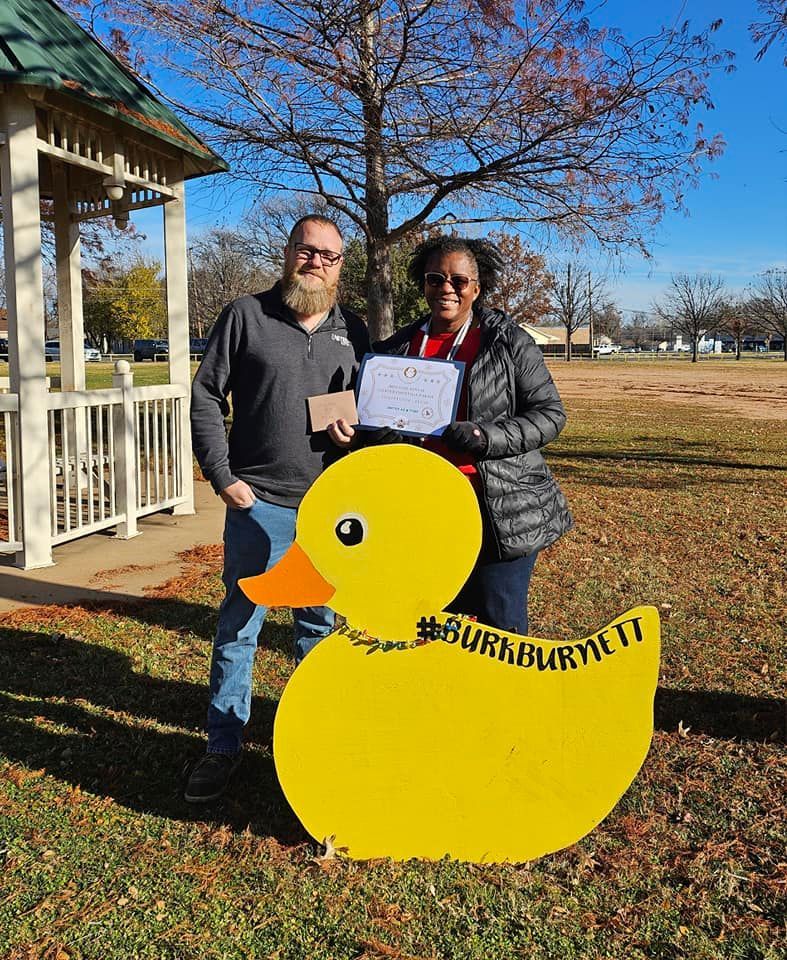 Two people stand next to a large yellow duck cutout holding a certificate outside near a gazebo.
