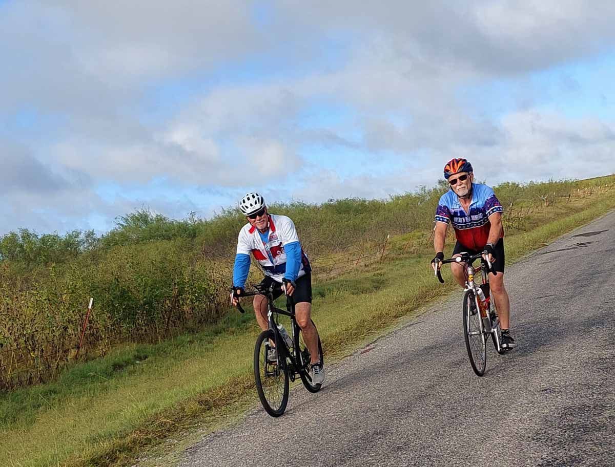Two cyclists ride uphill on a paved road, smiling, under a partly cloudy sky.