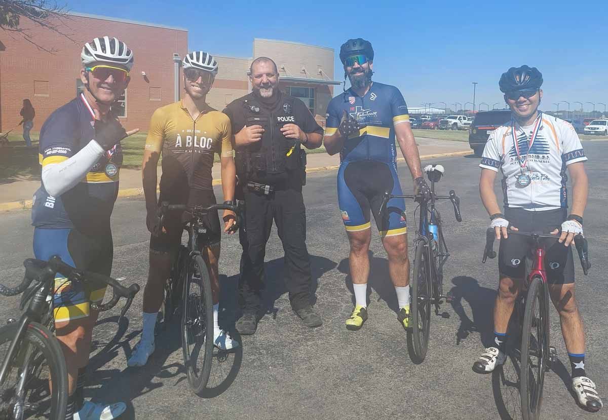 Five cyclists pose with a police officer outdoors; sunny day, wearing helmets, holding bikes.