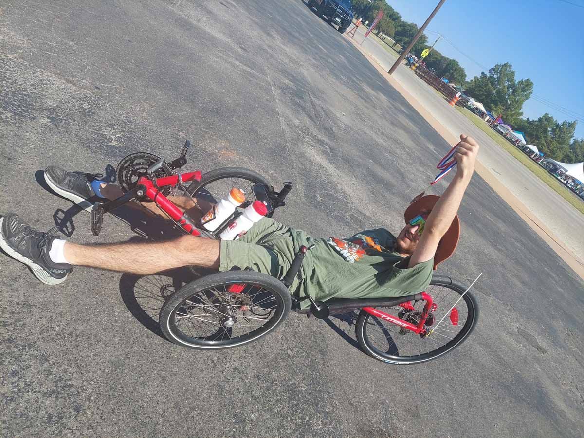 Man on red recumbent bike holding a drink outdoors on pavement.