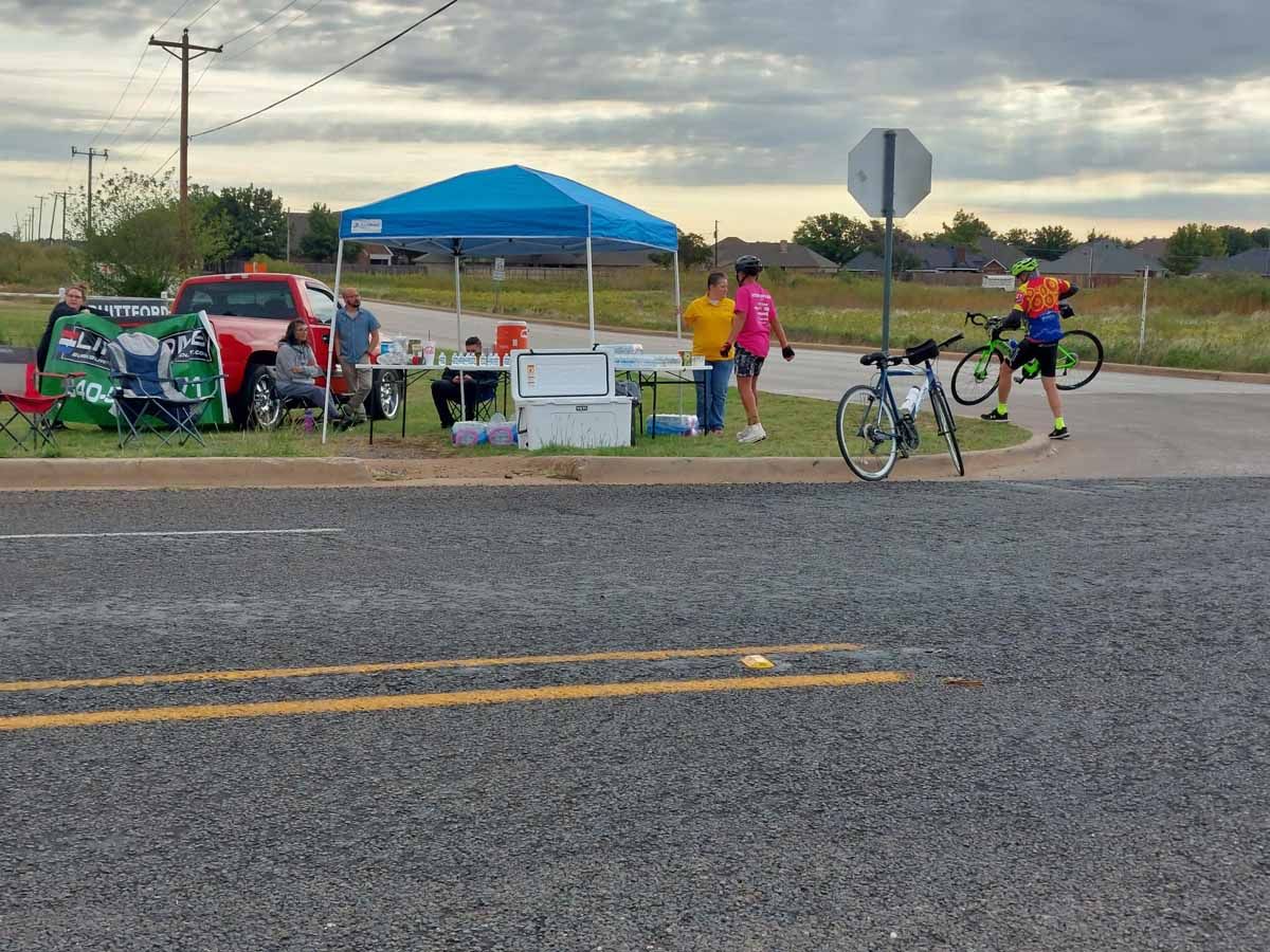 Cyclists at rest stop with canopy, red car, volunteers, bikes, on asphalt road.
